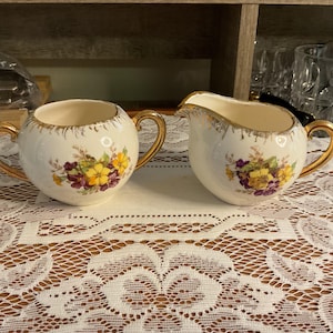 May include: A cream-colored sugar bowl and creamer set with gold handles and trim. The set is decorated with a floral pattern featuring yellow and purple flowers. The sugar bowl is round, and the creamer has a spout.