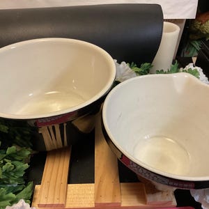 May include: Two white ceramic mixing bowls with black rims. The bowls are sitting on a wooden surface.