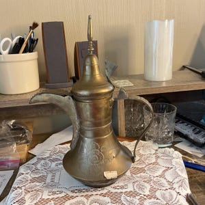 Puede incluir: Una cafetera de latón vintage con un pico largo, un asa ornamentada y un diseño floral. La olla tiene una tapa alta y puntiaguda y una pequeña etiqueta adjunta. Se encuentra sobre un mantel de encaje blanco.