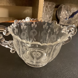 May include: A clear glass sugar bowl with two handles and a scalloped rim. The bowl features etched floral designs, including flowers and leaves. The bowl is likely vintage and adds a touch of elegance to any table setting.