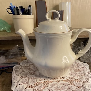 May include: Cream-colored ceramic teapot with a lid and handle. The teapot has a curved spout and a gold-colored trim. It sits on a white lace tablecloth. A matching lid with a loop handle is on top.