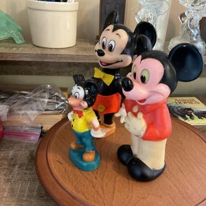 May include: Three vintage Mickey Mouse figurines. The tallest Mickey wears a red jacket, black shorts, and black shoes. The middle Mickey has a yellow bow tie, orange shorts, and black shoes. The smallest Mickey wears a yellow shirt and blue pants.