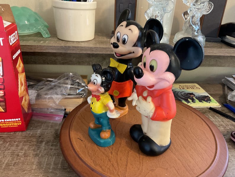 May include: Three vintage Mickey Mouse figurines. The tallest Mickey wears a red jacket, black shorts, and black shoes. The middle Mickey has a yellow bow tie, orange shorts, and black shoes. The smallest Mickey wears a yellow shirt and blue pants.