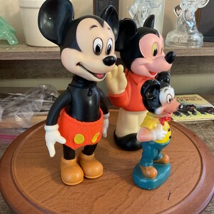 May include: Three vintage Mickey Mouse figurines. The largest Mickey is in a classic pose with orange shorts and yellow buttons. The middle Mickey is waving, wearing an orange jacket. The smallest Mickey is in a yellow shirt and blue pants.