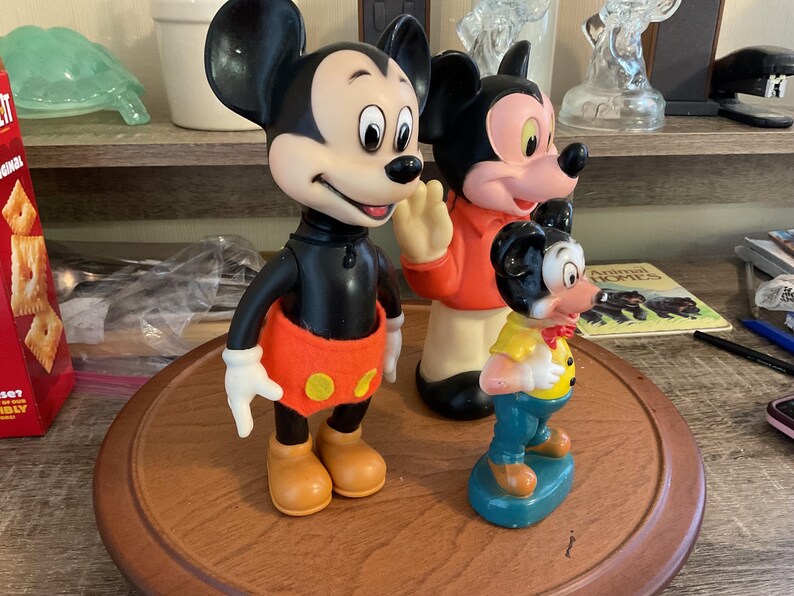 May include: Three vintage Mickey Mouse figurines. The largest Mickey is in a classic pose with orange shorts and yellow buttons. The middle Mickey is waving, wearing an orange jacket. The smallest Mickey is in a yellow shirt and blue pants.