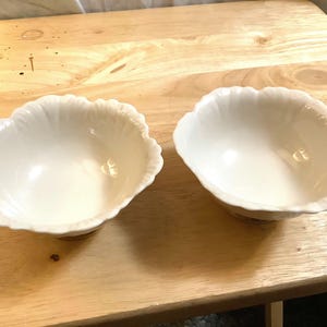 May include: Two off-white, scalloped-edge bowls. The bowls have a petal-like design around the rim and sit on a small, round base. The interior of the bowls is smooth and glossy, reflecting the light. They are placed on a light-colored wooden surface.