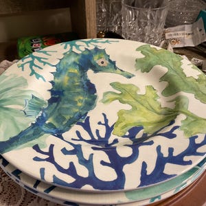 Könnte beinhalten: Ein Stapel weißer Keramikteller mit einem Meeresleben-Design. Jeder Teller zeigt ein Aquarell-Seepferdchen in Blau- und Grüntönen, umgeben von blauen Korallen und grünen Algen.