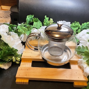 May include: A clear glass teapot with a stainless steel lid and strainer. The teapot is sitting on a wooden stand.