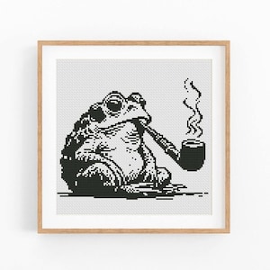Puede incluir: Impresión artística en punto de cruz en blanco y negro de una rana fumando en pipa. La rana está sentada, con la pipa en la boca y humo saliendo de la pipa. La obra de arte está enmarcada en un marco de madera marrón claro.