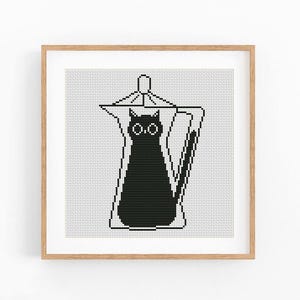 Puede incluir: Un patrón de punto de cruz enmarcado de forma cuadrada que presenta la silueta de un gato negro dentro del contorno de una cafetera italiana. El diseño utiliza hilo negro sobre tela de rejilla blanca, en un marco de madera clara.