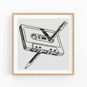 May include: Black and white cross-stitch art print of a cassette tape with a pencil inserted through it. The artwork is framed in a light brown wooden frame and set against a white background.