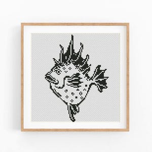 May include: A square framed cross stitch pattern featuring a black and white graphic of a spiky fish. The design is composed of small, distinct grid-like stitches on a white background, enclosed within a light-colored wooden frame.