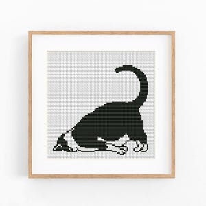 Peut inclure: Un motif de point de croix carré représentant un chat noir et blanc dans une position de jeu avec la queue arquée vers le haut. Le design est présenté dans un cadre en bois clair, mettant en valeur une esthétique pixelisée sur une grille blanche.