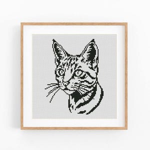 Può includere: Uno schema a punto croce quadrato incorniciato che presenta un'illustrazione grafica in bianco e nero di una testa di gatto. Il disegno è composto da una griglia di piccoli punti su sfondo bianco, racchiuso in una cornice di legno chiaro.