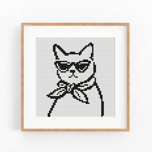 以下が含まれることがあります： サングラスとスカーフをつけた猫のクロスステッチアート。猫は白黒で、正方形のグリッドの背景があります。アートワークは、家の装飾に最適な、明るい茶色の木製フレームに額装されています。