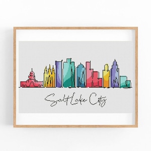 May include: A framed print featuring a colorful Salt Lake City skyline. The artwork showcases a line drawing of the city's buildings in various colors, including red, yellow, teal, and purple, with the city name in cursive.