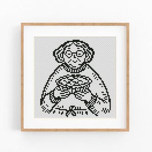 Può includere: Uno schema a punto croce incorniciato che presenta un'illustrazione pixelata in bianco e nero di una persona con capelli ricci e occhiali, che indossa un maglione e tiene in mano un oggetto circolare, in una cornice quadrata in legno chiaro.