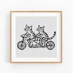 以下が含まれることがあります： タンデム自転車に乗った2匹の猫のクロスステッチアート。猫と自転車は白黒です。このアートワークは、明るい茶色の木製フレームに収められており、家の装飾に最適です。