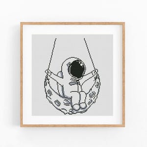 May include: A framed cross-stitch art piece depicts an astronaut in a spacesuit swinging on a crescent moon. The astronaut is in a seated position with arms outstretched. The artwork is in black and white, with a light gray background and a wooden frame.