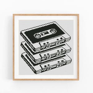 May include: A framed, pixelated art print featuring a stack of three black and white cassette tapes. The artwork has a retro, 80s or 90s aesthetic, with a simple, graphic design. The tapes are detailed with reel and label features.