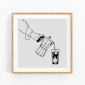 May include: Black and white line art print of a hand pouring coffee from a stovetop espresso maker into a glass. The artwork is framed in a light brown wooden frame, set against a white background.