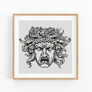 May include: A framed black and white cross-stitch of a classical Greek mask with an angry expression. The mask has a detailed headdress and flowing hair. The artwork is displayed in a light brown wooden frame.