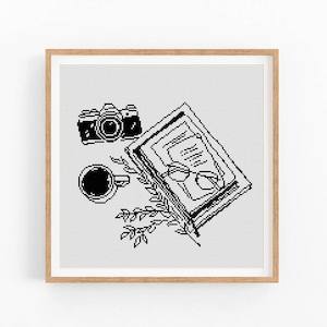 May include: A square cross-stitch pattern in a light wood frame featuring a black and white pixelated design of a camera, an open book with glasses resting on the pages, a cup of coffee, and a small leafy branch on a white grid background.