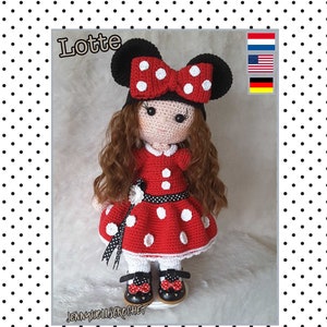 Puede incluir: Una muñeca de crochet que lleva un vestido de lunares rojos y blancos con un lazo negro y una diadema de lunares negros y blancos. La muñeca tiene el pelo castaño y los ojos marrones. La muñeca está de pie sobre una superficie blanca. El texto "Lotte" es visible en la imagen.