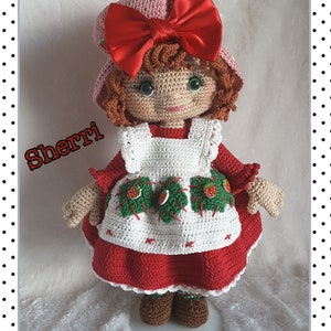 May include: A crocheted doll wearing a red dress with a white apron and a pink hat with a red bow. The doll has brown hair and green eyes. The doll is standing on a white surface. The text "Sherri" is written in black letters on the left side of the image. The text "Adrienne" is written in black letters on the bottom left side of the image. The text "PIC-COLLAGE" is written in black letters on the bottom right side of the image.