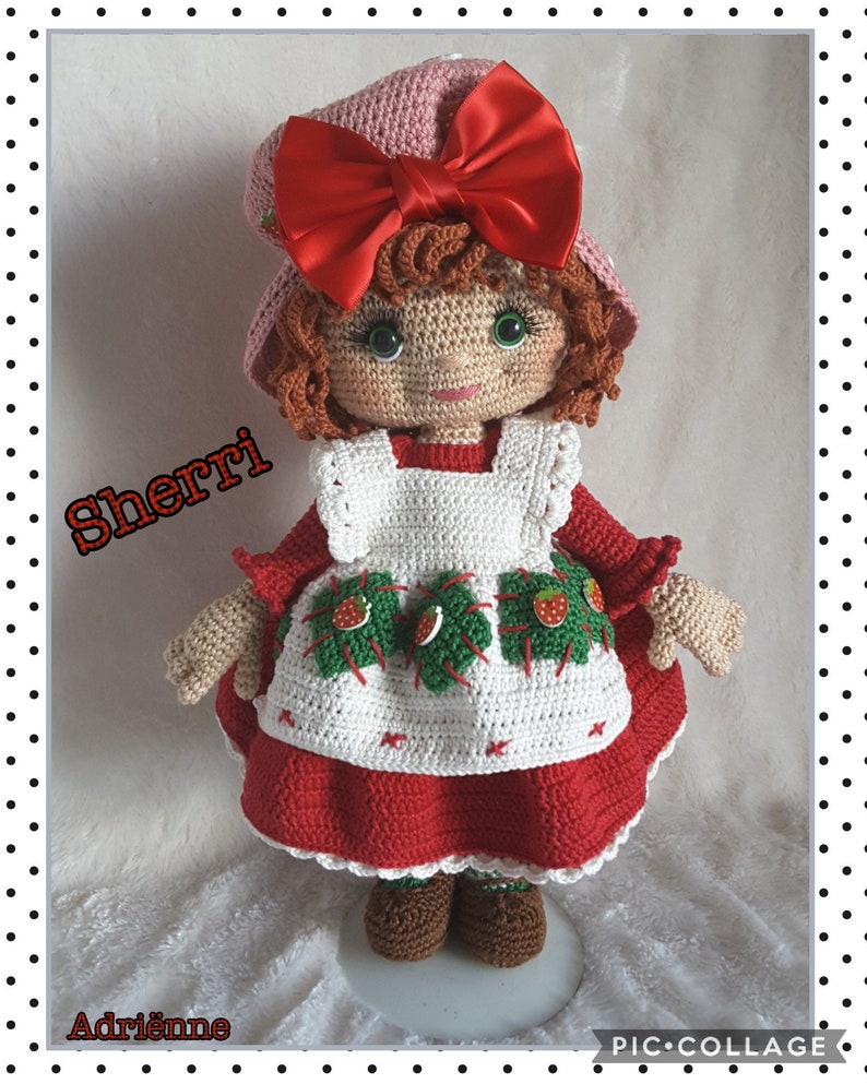 May include: A crocheted doll wearing a red dress with a white apron and a pink hat with a red bow. The doll has brown hair and green eyes. The doll is standing on a white surface. The text "Sherri" is written in black letters on the left side of the image. The text "Adrienne" is written in black letters on the bottom left side of the image. The text "PIC-COLLAGE" is written in black letters on the bottom right side of the image.