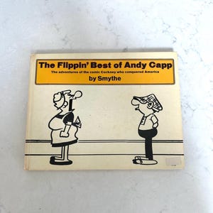 May include: A yellow and black book cover with the title "The Flippin' Best of Andy Capp" and the subtitle "The adventures of the comic Cockney who conquered America by Smythe".  Two black and white cartoon characters are shown on the cover.