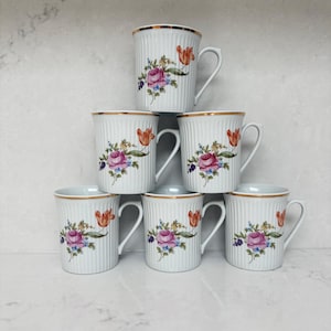 Peut inclure: Un ensemble de sept tasses en céramique blanche avec des détails côtelés verticaux. Chaque tasse présente un motif floral avec des roses roses, des tulipes oranges et des accents bleus. Les bords sont dorés.