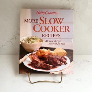 May include: A cookbook titled "More Slow Cooker Recipes" by Betty Crocker, featuring a photo of a meal with ribs, mashed potatoes, and cornbread. The book is displayed on a gold-colored metal stand.