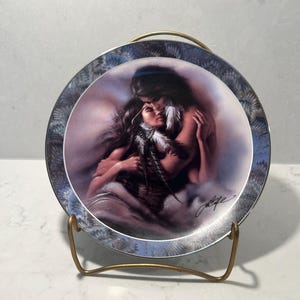 May include: A decorative plate featuring a romantic image of two people embracing, with long dark hair and feathers. The plate has a silver and blue patterned border and is displayed on a gold-colored metal stand.