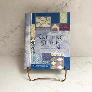 May include: A book titled "The Knitting Stitch Bible" is displayed in a gold-coloured metal stand. The book has a blue cover with various knitting stitch patterns and the author's name, Maria Parry-Jones.
