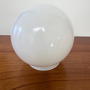 May include: A spherical white glass lamp shade with a smooth, glossy finish. The globe features a circular opening at the base with a slightly flared rim, designed for attachment to a light fixture. The surface is opaque and reflects light.