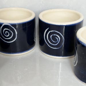 May include: Three cylindrical ceramic napkin rings featuring a deep navy blue glazed exterior with a hand-painted white spiral design. Each piece has a smooth, cream-colored interior rim and a glossy finish, showcasing a simple, uniform shape.