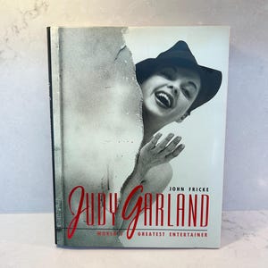 May include: A hardcover book titled Judy Garland: World's Greatest Entertainer by John Fricke. The cover features a black and white photograph of Judy Garland wearing a hat, peeking from behind a torn paper edge, with red text at the bottom.