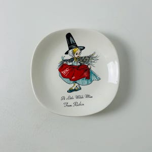 May include: A white ceramic dish featuring a colourful illustration of a girl in a red dress and a black hat. The text "A Little Welsh Miss From Ruthin" is printed on the dish.