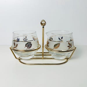 May include: A gold metal stand with two glass creamer and sugar containers. The containers have a frosted white background with a gold band and a leaf design in shades of brown, gold, and blue.