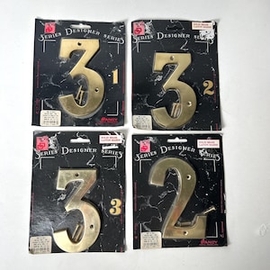 May include: Four gold-colored metal house numbers, 3 and 2, packaged in individual black and gold cardboards with the text "Series Designer Series" and "Solid Brass Laiton Massif".