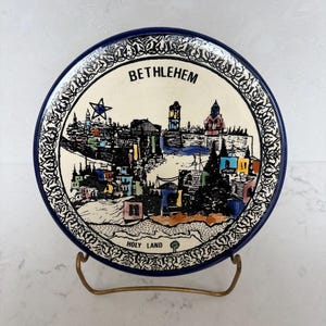 May include: A round ceramic decorative plate featuring a hand-painted illustration of the Bethlehem skyline. The design includes the text BETHLEHEM and HOLY LAND, a blue star, and colorful building accents, all framed by a black and white patterned border.