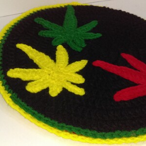 Könnte beinhalten: Ein rundes, schwarzes gehäkeltes Objekt mit drei Cannabisblatt-Designs in Gelb, Grün und Rot. Der Rand ist mit gelbem, grünem und gelbem Garn verziert.