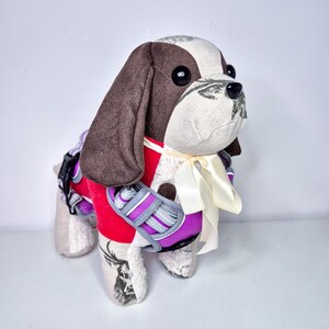 May include: A soft toy dog with brown and white fur, large brown ears, and black button eyes. The dog wears a red vest with a purple and grey harness and a cream ribbon. The dog's legs have a printed design.