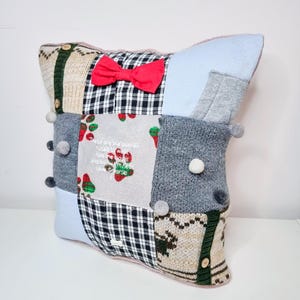 May include: A patchwork cushion with a red bow, made from different types of knit fabric. The cushion has a light blue background and features a variety of colours and patterns, including plaid, stripes, and polka dots. The cushion is decorated with white pom-poms and has an embroidered message that reads: "These are the paw prints of love, we walk together and leave our memories in the sand."