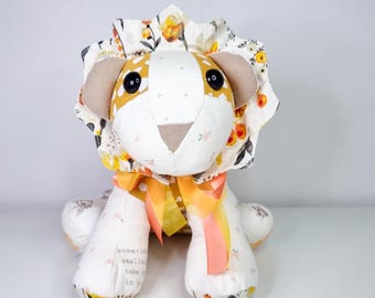 May include: A white and yellow fabric lion with a floral print on its head. The lion has black eyes and a pink ribbon around its neck. The lion is sitting on a white surface. The text "sometimes smaller things take up more room in your heart" is printed on the lion's leg.