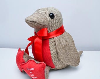 May include: A stuffed bird with a brown tweed body, black eyes, and a red satin ribbon around its neck. The bird's feet are red, with the words "Forever in our hearts" embroidered on one foot. A decorative item.