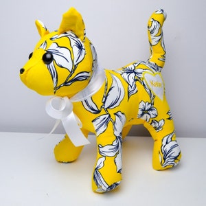 May include: A yellow and white floral fabric cat plush toy with a white ribbon around its neck. The toy has black eyes and a white heart-shaped patch on its back that says "Katie".