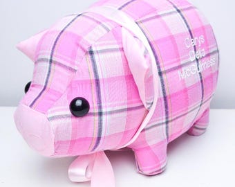 May include: A pink plaid piggy bank with black eyes and a pink snout. The pig has a pink ribbon around its neck and the names "Carys Delia McGuinness" are embroidered on its side. A decorative item.