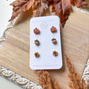 May include: A set of three pairs of stud earrings on a white display card labeled Kalico Citten Designs. The earrings feature miniature clay shapes: orange pumpkins with green stems, brown acorns with tan caps, and yellow sunflowers with brown centers.